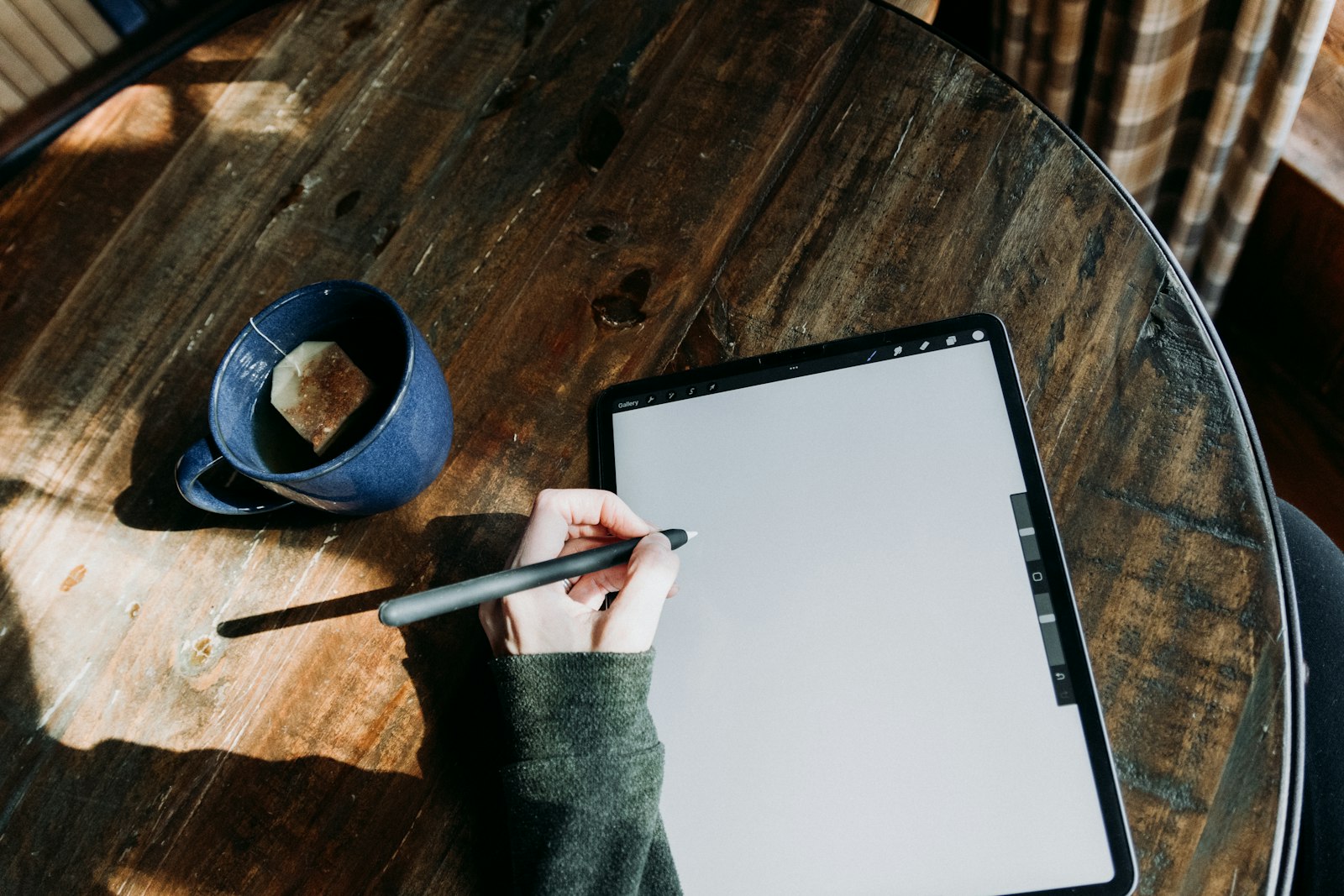 A person at a wooden table writing with a stylus on a tablet beside a mug of coffee