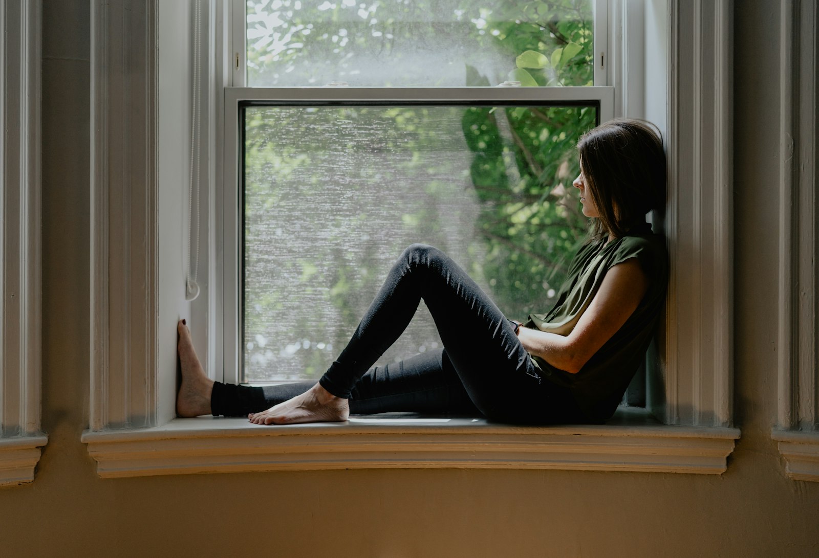 A person sitting quietly on a wide window sill, looking out at green trees
